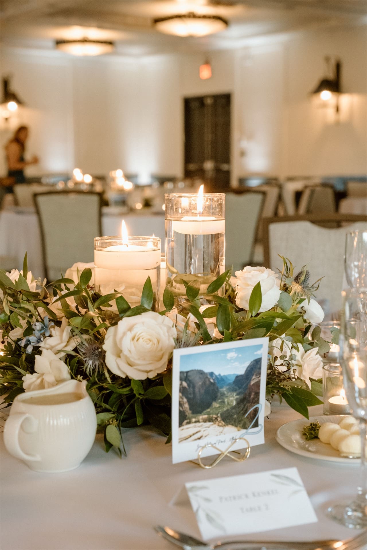 Floral Wedding Centerpiece with roses, leaves, and water glasses topped with candles. In front of centerpiece is a polaroid picture of mountain tops from wedding couple's hiking trip.