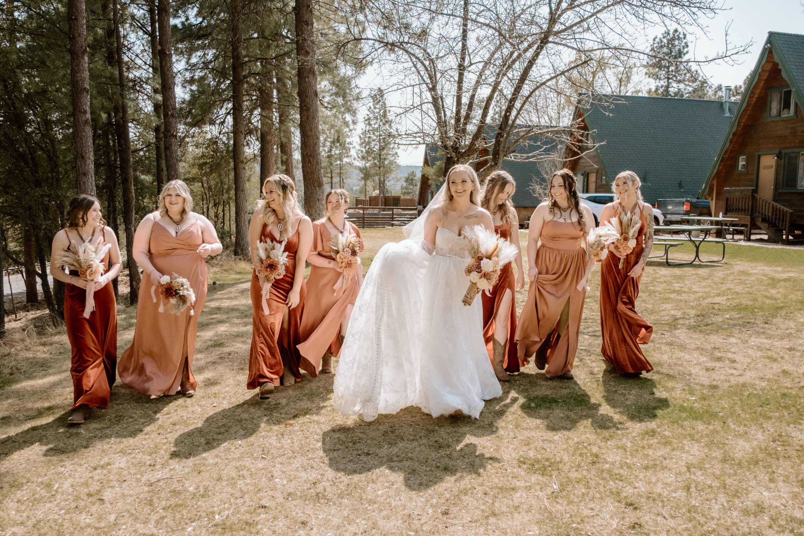 Bride walking down forest field with neighboring cabins, laughing alongside seven bridesmaids in peach colored gowns.