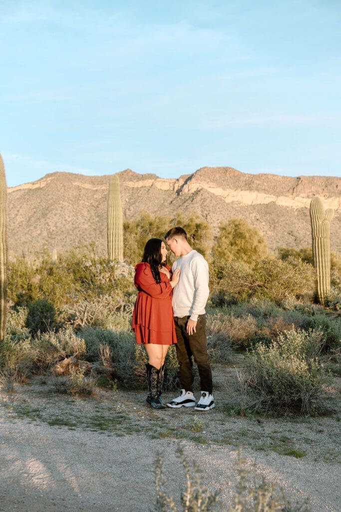 Couple posed with woman's hand placed on man's chest standing in front of Arizona desert mountains surrounded by cacti and natural bushes as landscape.